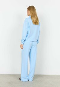 Frau mit langem blondem Haar, die steht und einen lockeren hellblauen Pullover sowie weit geschnittene Hosen trägt, mit dem Rücken zu einer weißen Wand.