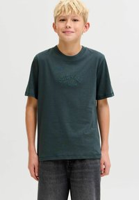 Camiseta de algodón verde oscuro con mangas cortas, cuello redondo y texto bordado "ORIGINALS" en el frente. Combinada con pantalones cortos de mezclilla grises.