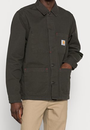 Mężczyzna ubranego w ciemnozieloną kurtkę zapinaną na guziki z trzema kieszeniami i beżowe spodnie, z ręką w prawej kieszeni kurtki.