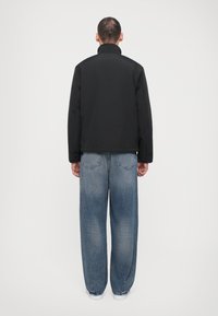 Svart jacka med hög krage och långa ärmar, bärs över löst sittande ljusblå jeans med blekt mönster och bakfickor.