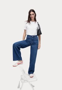 Højtaljede, brede denimjeans i blå, hvid t-shirt og sneakers med røde detaljer. Modellen står på en taburet og holder en sort håndtaske.