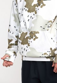 Sudadera con capucha en un patrón de camuflaje blanco, beige y oliva. Cuenta con un bolsillo tipo canguro y agujeros para los pulgares en los puños. Textura de tela suave.