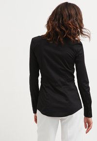 Czarna dopasowana koszula z długimi rękawami, wykonana z gładkiego materiału, z zapięciem na guziki przy mankietach i lekko zaokrąglonym dołem, noszona na białych spodniach.