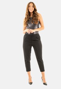 Pantalones negros ajustados con pierna cónica, combinados con una blusa sin mangas de color gris metálico. La modelo está de pie sobre tacones negros, con las manos unidas.