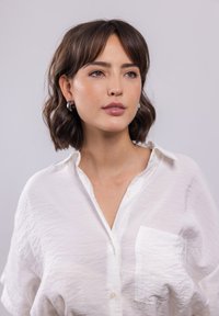 Weißes, lässig geschnittenes Hemd mit Knöpfen; verfügt über eine Brusttasche und eine leicht zerknitterte Textur. Das Modell trägt runde schwarze Ohrringe.