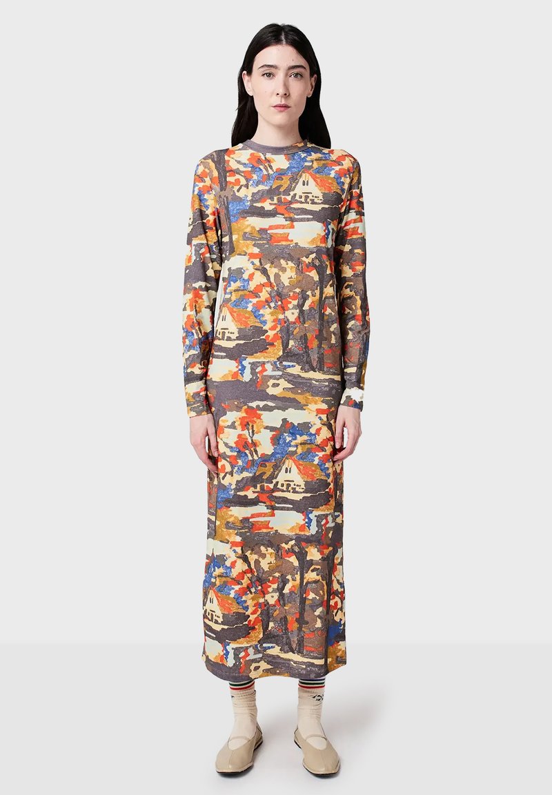 Woman wearing a long-sleeve, ankle-length dress with an abstract, multicolor pattern, beige shoes, and cream socks standing against a plain background.