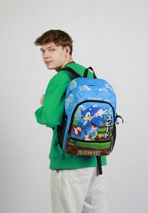 Mochila azul con un gráfico de un erizo azul con acentos verdes, nubes y una base marrón. Cintas ajustables y bolsillos con cremallera visibles.