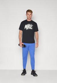 Musta Nike T-paita valkoisella logolla, siniset urheiluhousut ja mustat lenkkarit. Malli pitää mustaa vesipulloa ja seisoo neutraalilla taustalla.
