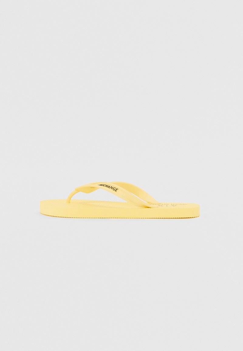 Gul flip-flop laget av gummi med flat såle, har teksturert fotseng og fleksible stropper. Logo synlig på stroppen.