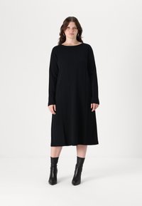 Abito nero a maniche lunghe con scollatura tonda, vestibilità ampia e cuciture verticali, indossato con stivali neri alla caviglia. Tessuto dalla consistenza liscia.