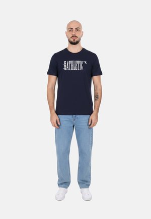 Uomo con barba e tatuaggi che indossa una t-shirt blu navy "Athletic", jeans azzurri chiari e sneakers bianche, in piedi davanti a uno sfondo bianco semplice.
