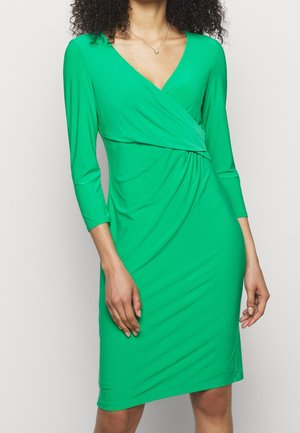 Kobieta w dopasowanej, zielonej do kolan sukience o stylu kopertowym z dekoltem w kształcie litery V i rękawami długości do połowy przedramienia.