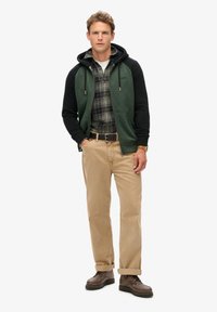 Sudadera verde y negra sobre una camisa de cuadros, combinada con pantalones beige y botas marrones. Los detalles notables incluyen un cierre frontal y un diseño de bolsillos.