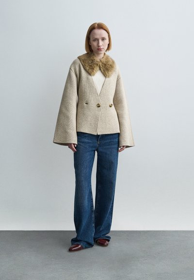 Μπεζ σακάκι από μείγμα μαλλιού με γιακά από ψεύτικη γούνα, διπλό πέτο, φαρδιά μανίκια· σετ με μπλε ψηλόμεσα τζιν και καφέ παπούτσια.