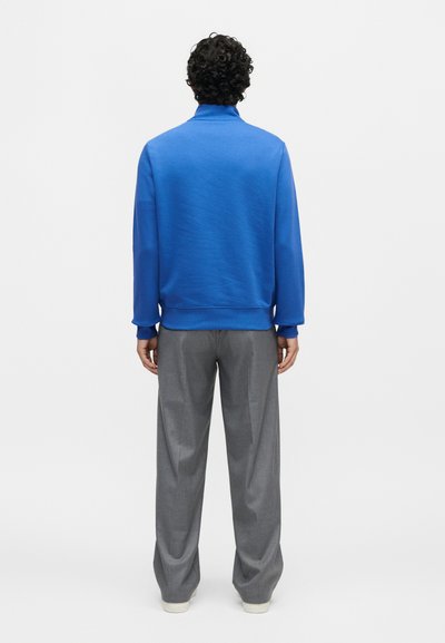 Άνδρας με σγουρά μαλλιά στέκεται με την πλάτη γυρισμένη, φοράει μια μπλούζα μακρυμάνικη σε βασιλικό μπλε και χαλαρό γκρι παντελόνι με λευκά παπούτσια σε λευκό φόντο.