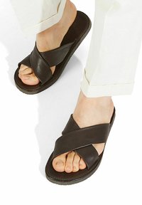 Sandali in pelle marrone con un design a fascetta incrociata e suola piatta. La superficie testurizzata e i dettagli delle cuciture arricchiscono l'aspetto complessivo.