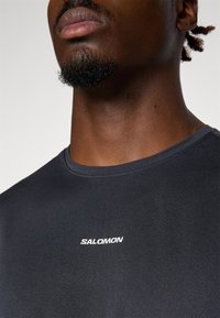 Schwarzes, strukturiertes Sportoberteil mit Rundhalsausschnitt, auf der Vorderseite mit einem weißen "SALOMON"-Logo versehen. Nahaufnahme des Ausschnitts und des oberen Brustbereichs.