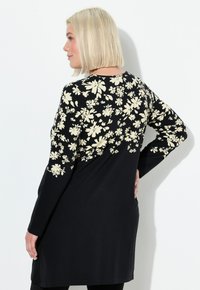Vrouw met kort blond haar, gekleed in een zwarte jurk met lange mouwen en een crème bloemenpatroon op de bovenrug en mouwen, staande in profiel.