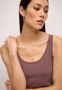 Halenka bez rukávů s lodičkovým výstřihem v jemné mauve barvě. Hladká textura a přiléhavý střih. Modelka pózuje s jednou rukou na rameni.