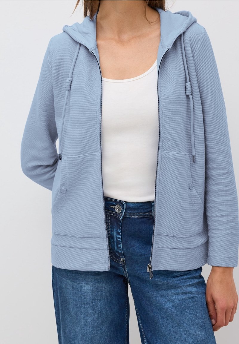 Femme portant un sweat à capuche bleu clair zippé sur un débardeur blanc et un jean bleu, debout devant un fond uni.