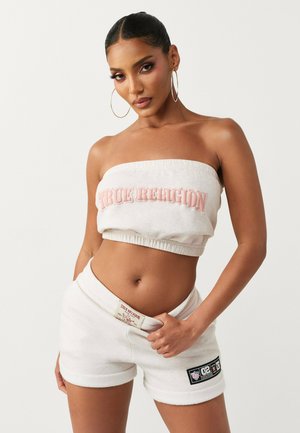 Vrouw met grote oorringen, gekleed in een witte tube top en bijpassende short met "True Religion" branding, poserend tegen een effen achtergrond.