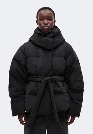 Personne portant une veste matelassée noire avec un col montant et une ceinture nouée à la taille, debout devant un fond clair uni.