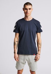 Mörkblå bomulls-t-shirt med kort ärm, med vita chevronränder på axlarna och en liten röd etikett vid fållen.