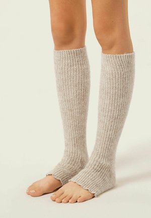 Kniehoge beenwarmers in zacht beige breiwerk, met een geribbelde structuur en een golvende zoom. Open teenontwerp laat blote voeten zien.