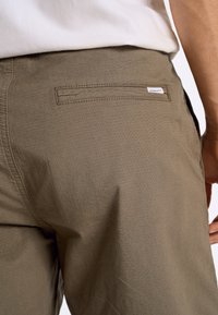 Pantaloni verde oliva con un tessuto testurizzato, dotati di una tasca posteriore con etichetta e dal design a vestibilità dritta. Le cuciture sono discrete.