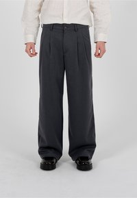 Homme portant un pantalon large gris foncé, des chaussures noires et une chemise à manches longues blanche à rayures, debout devant un fond uni.