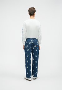 Oseba v beli dolgi srajci z dolgimi rokavi in modrih kavbojkah, okrašenih z belimi simboli yin-yang, stoji s hrbtom obrnjenim proti beli ozadju.