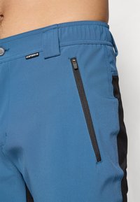 Plave kratke hlače s crnim bočnim detaljima, koje imaju džep s patentnim zatvaračem, glatku teksturu i fleksibilni pojas. Oznaka loga vidljiva na pojasu.