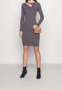 Robe longue à manches longues grise côtelée avec un détail découpé, associée à un petit sac à main beige et des bottines noires. Tissu texturé, silhouette ajustée.