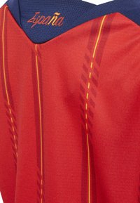 Červený a námornícky športový dres so žltými vertikálnymi pruhmi a vyšitým nápisom "España" na námorníckom golieri v oranžovej farbe.