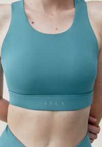 Urheiluliivit teal-värisestä kankaasta, jossa on sileä pinta. Ominaisuuksiin kuuluu leveä vyötärö, jossa on logo, ja urheilullinen selkäosa, joka tarjoaa tukea aktiviteettien aikana.