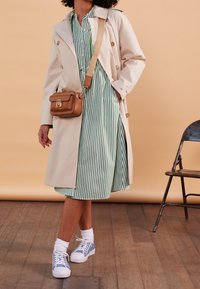 Trench coat beige con bottoni dorati sopra un vestito a strisce verdi e bianche. Borsa a tracolla marrone con logo, calze bianche e sneakers blu.