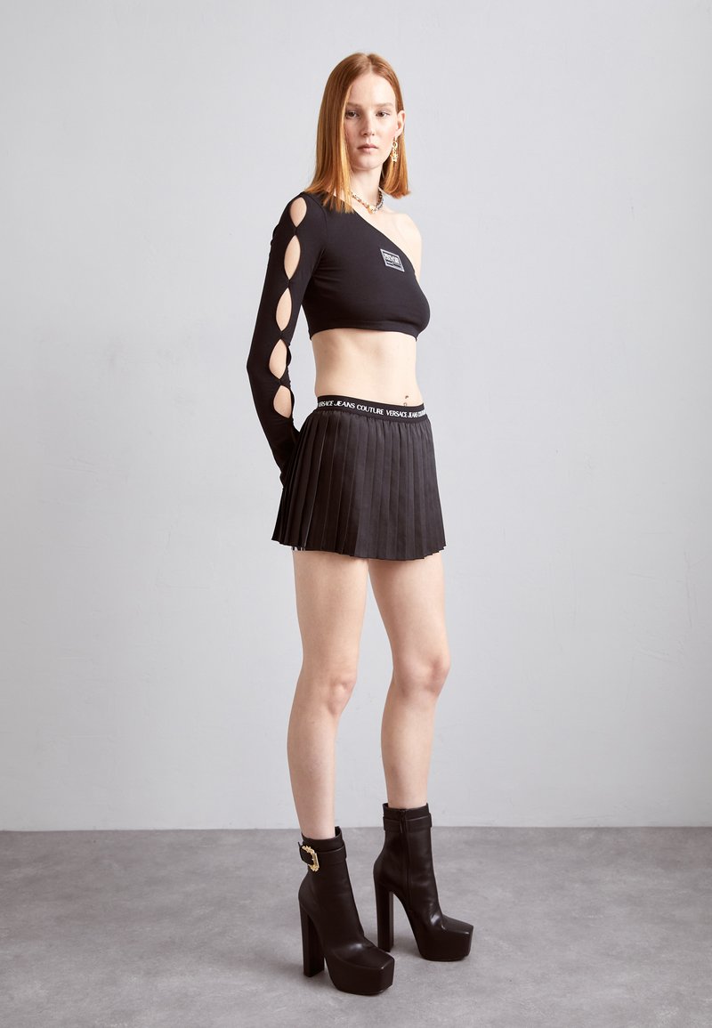 Top nero senza spalle con dettaglio a foro su una manica, abbinato a una mini gonna nera plissettata e stivaletti neri con tacco largo, con accenti dorati.