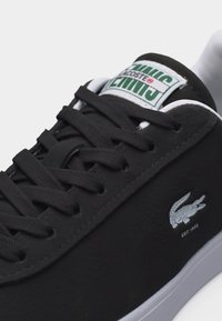 Μαύρο αθλητικό παπούτσι τένις Lacoste με μαύρα κορδόνια, λευκή σόλα και λευκό λογότυπο κροκόδειλου στο πλάι, με το όνομα της μάρκας στην ετικέτα της γλώσσας.