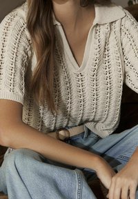 Pull-over en tricot couleur crème clair avec une texture côtelée, manches courtes et col en V, accompagné d'une ceinture beige et d'un jean bleu.