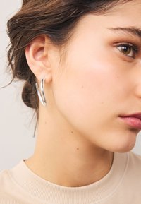 Silver hoop earrings with a smooth, shiny surface, featuring a curved design. The model has dark hair and wears a beige top.