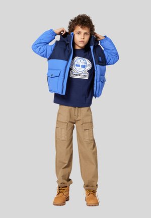 Doudoune bleue et marine avec capuche, portée sur un t-shirt à logo Timberland marin. Un pantalon cargo kaki et des bottes de travail beiges complètent la tenue.