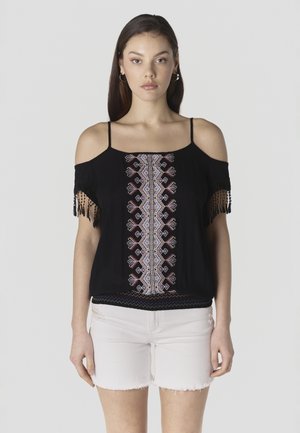 Mujer con blusa negra de hombros descubiertos con bordado geométrico y mangas con flecos, combinada con shorts de mezclilla blanca desgastada, de pie derecho.