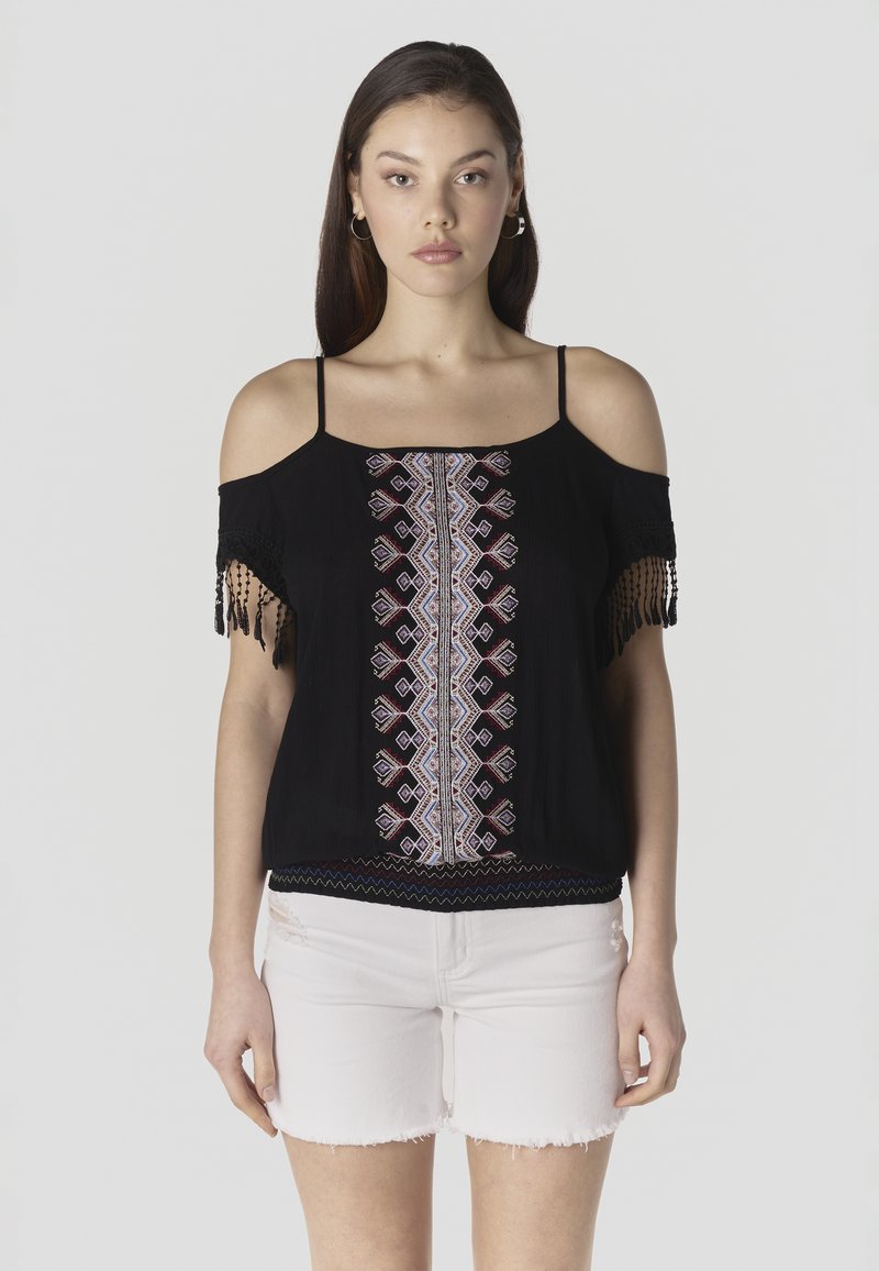 Mujer con blusa negra de hombros descubiertos con bordado geométrico y mangas con flecos, combinada con shorts de mezclilla blanca desgastada, de pie derecho.