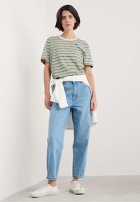 T-shirt w paski w kolorach żółtym, niebieskim i białym z krótkimi rękawami, w połączeniu z jasnoniebieskimi dżinsami i białymi trampkami, z białym swetrem związanym w talii.