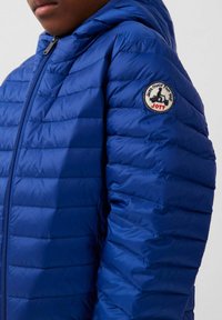 Blauwe pufferjas met een doorgestikt ontwerp, voorzien van een capuchon, ritssluiting en een ronde logopatch op de linkerschouder. Zachte textuur.