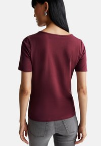 Burgundské tričko s krátkým rukávem vyrobené z žebrované tkaniny, s kulatým výstřihem a slim fit, v kombinaci se šedými džíny. Zobrazeno ze zadní strany.