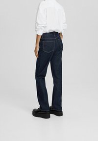 Jean de mezclilla de lavado oscuro con corte recto, que cuenta con dos bolsillos traseros y detalles de costura visibles. Combinado con una camisa blanca de manga larga.