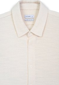 Polo shirt a maglia di colore crema, con texture, colletto standard, cinque bottoni e etichette che indicano la taglia e la vestibilità.