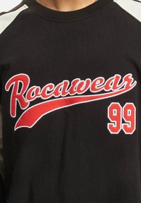 Svart ribbet genser med rød brodert "Rocawear" tekst og "99" på en hvit detalj, med kontrastfargede ermer.