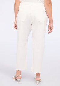Persona con pantalones cortos blancos y sandalias de tacón abiertas en la parte de atrás, de pie contra un fondo claro y liso.